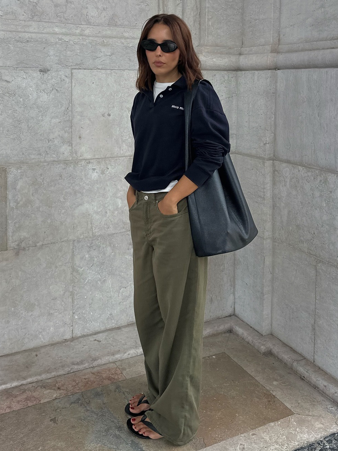 Style influencer Debora Rosa poses in a Miu Miu polo shirt layered over a white tee, a black leather tote bag, baggy green pants, and black flip-flop sandals.
