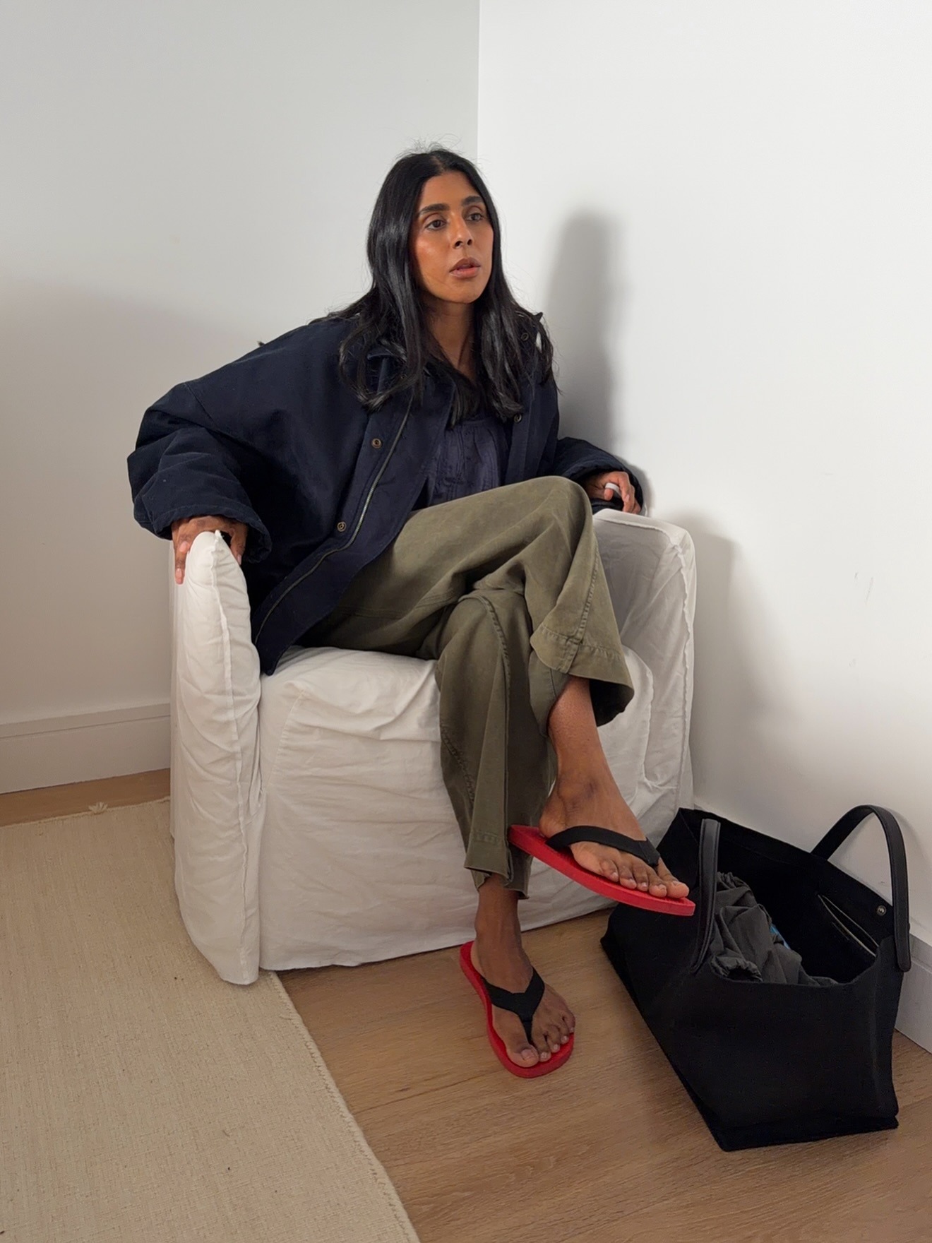 British style influencer Monikh Dale poses in a white slipcover chair wearing an oversize navy blue jacket baggy green pants, black suede tote bag, and black and red flip-flop sandals.