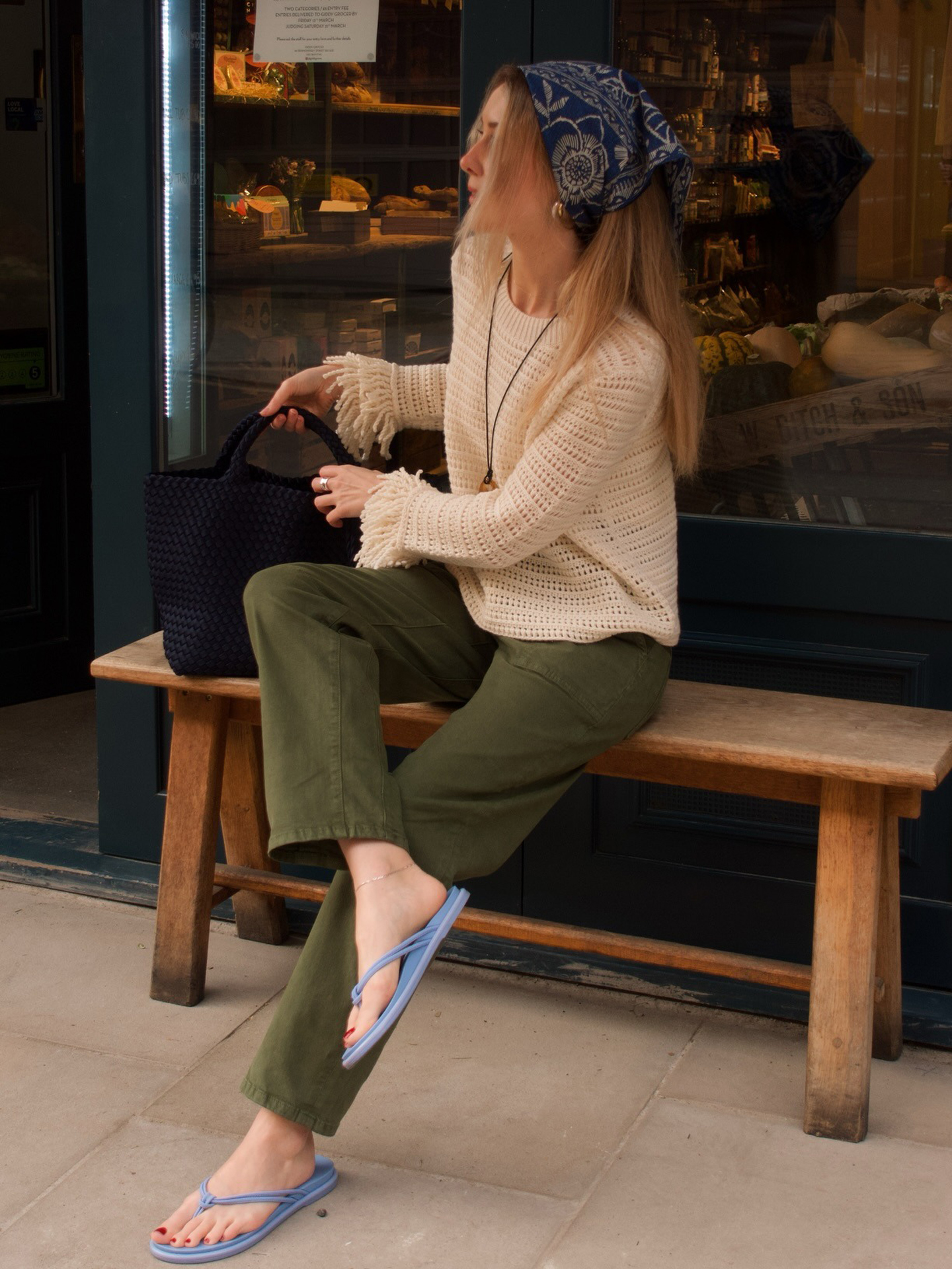 British style influencer poses on a bench outside a store in London wearing a blue printed bandana head scarf, an open-knit sweater, black tote bag, green pants, and light blue flip-flop sandals.
