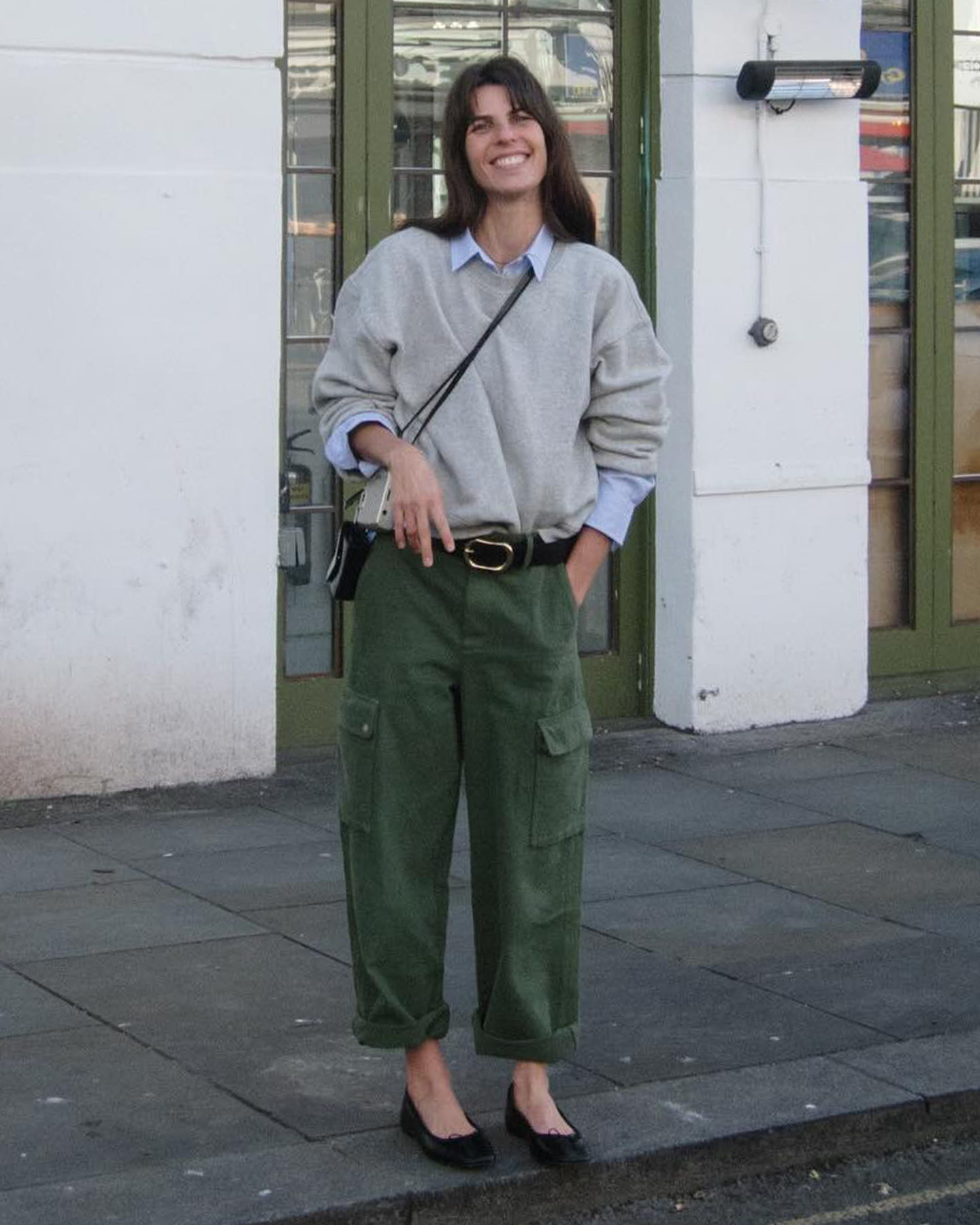 French style influencer Laura Vidrequin Roso poses in a gray sweatshirt layered over a blue button-down shirt, a black belt, crossbody bag, cuffed green cargo pants, and black flats.