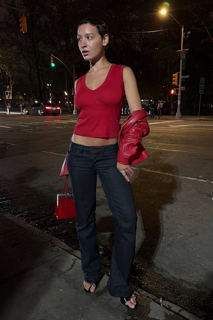 A woman wearing a red top, red leather jacket, dark wash jeans, and black sandals.