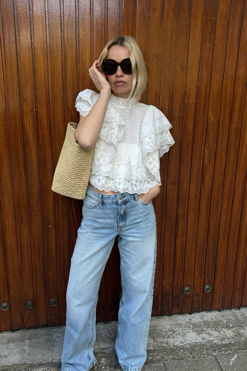 A woman wearing a lace white top, blue jeans, and a straw woven bag.
