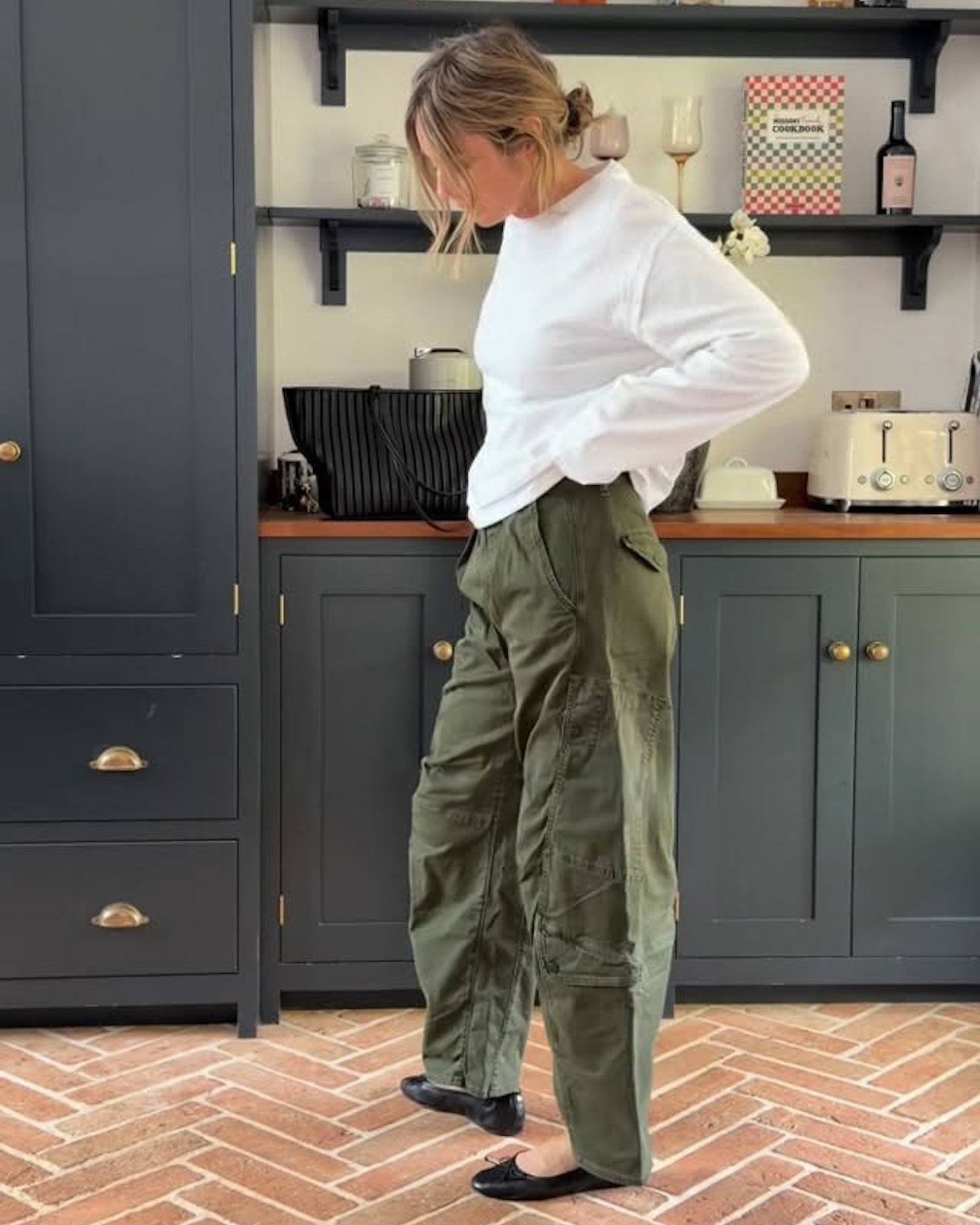British style influencer Emma Rose Thatcher poses in her kitchen wearing an oversize long-sleeve white T-shirt, a woven black bag, green barrel pants, and black ballet flats.