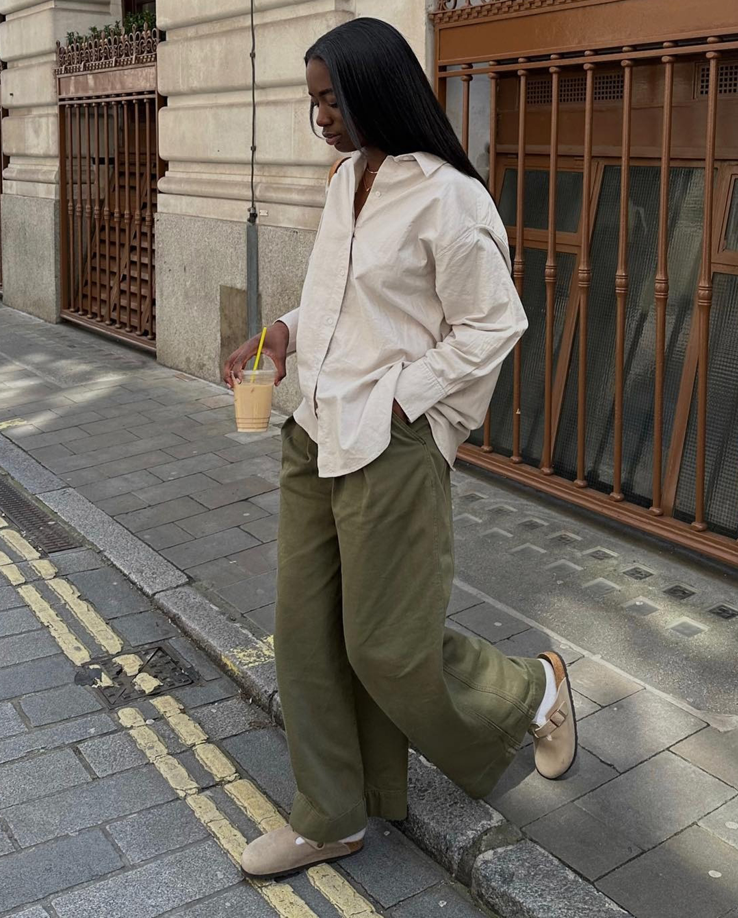 British style influencer Liv Madeline poses on a sidewalk wearing an oversize beige button-down shirt, wide-leg green pants, white socks, and tan Birkenstock Boston clogs.