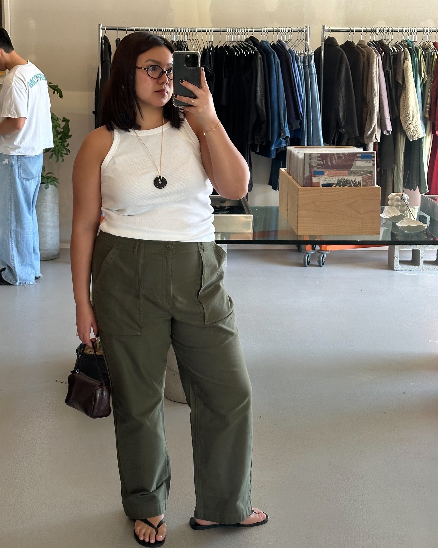 Style influencer Marina Torres poses for a mirror selfie wearing oval optical eye glasses a white tank top, cord pendant necklace, relaxed green pants, and black flip-flop sandals.